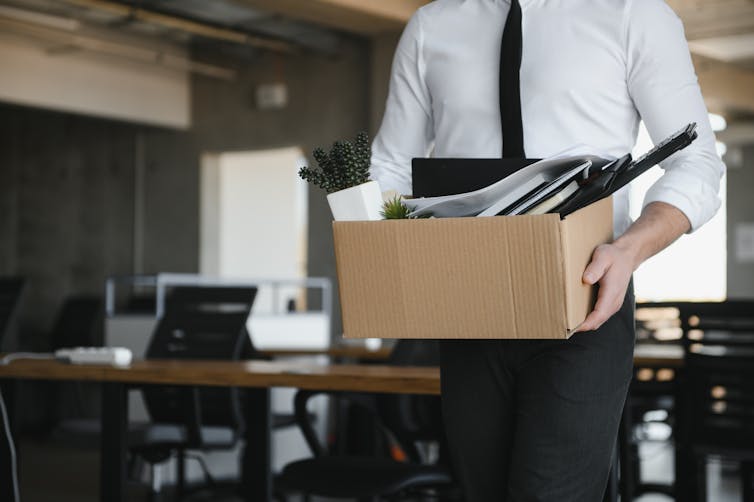 Person in a suit seen from the neck-down carrying a cardboard box full of office supplies