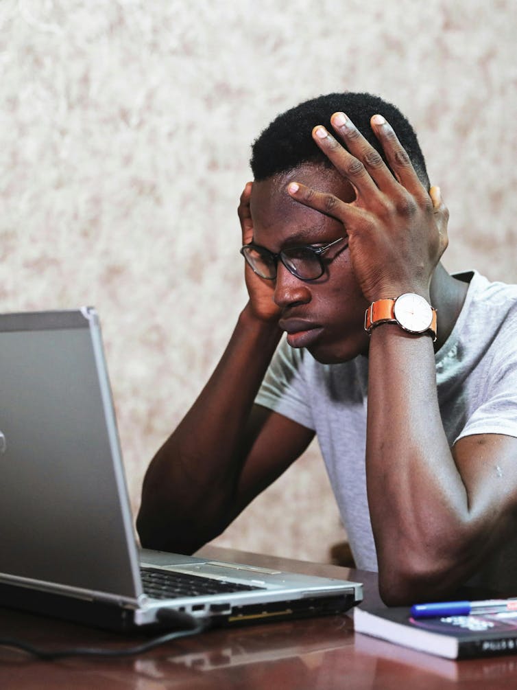 A man sits at a computer looking stressed, holding his head in his hands.